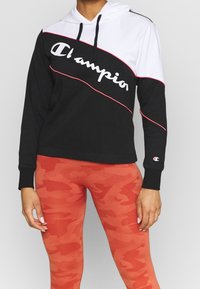 Svart och vit hoodie med röda detaljer och "Champion"-logotyp, kombinerad med orange kamouflageleggings. Tyget verkar mjukt med en bekväm passform.