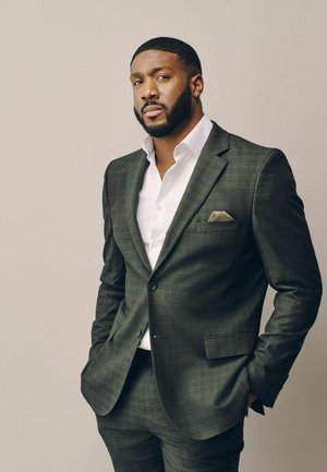 Man with short hair and beard wearing a dark green checkered suit with a white shirt and pocket square, standing with hands in pockets.