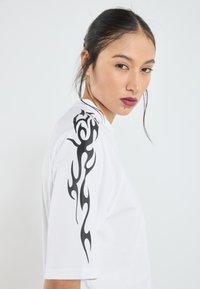Camiseta de algodón blanca con un diseño de llamas negras en el hombro izquierdo, de mangas cortas y un corte relajado. Textura suave.