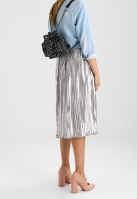 Jupe midi plissée argentée, associée à une chemise en denim claire, un sac seau noir texturé et des chaussures à talons carrés rose pâle.