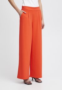 Wijdvallende broek in feloranje, gemaakt van een gladde stof, voorzien van een elastische tailleband, plooien en zijzakken, gecombineerd met zwarte hakken sandalen.