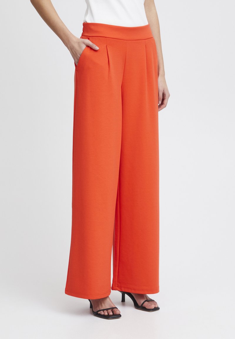 Wijdvallende broek in feloranje, gemaakt van een gladde stof, voorzien van een elastische tailleband, plooien en zijzakken, gecombineerd met zwarte hakken sandalen.