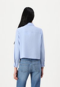 Lys blå skjorte med lange ermer og krage, med knappede mansjetter og en glatt tekstur. Kombinert med mellomblå jeans.