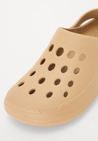 Light tan clogs made of flexible plastic with round ventilation holes, a smooth exterior, and textured sole for grip.