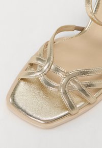 Gullmetalliske høyhælte sandaler med kryssende stropper og firkantet tå. Glatt, glitrende tekstur med en polstret beige såle.