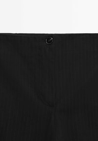 Ceinture en tissu noir texturé avec une fermeture à bouton noir unique et des lignes de couture verticales visibles.