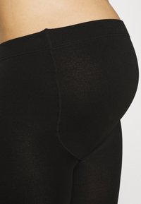 Zwarte leggings hebben een gladde, rekbare stof met een hoge tailleband. De textuur lijkt op sommige plaatsen iets doorschijnend, zonder zichtbare patronen.