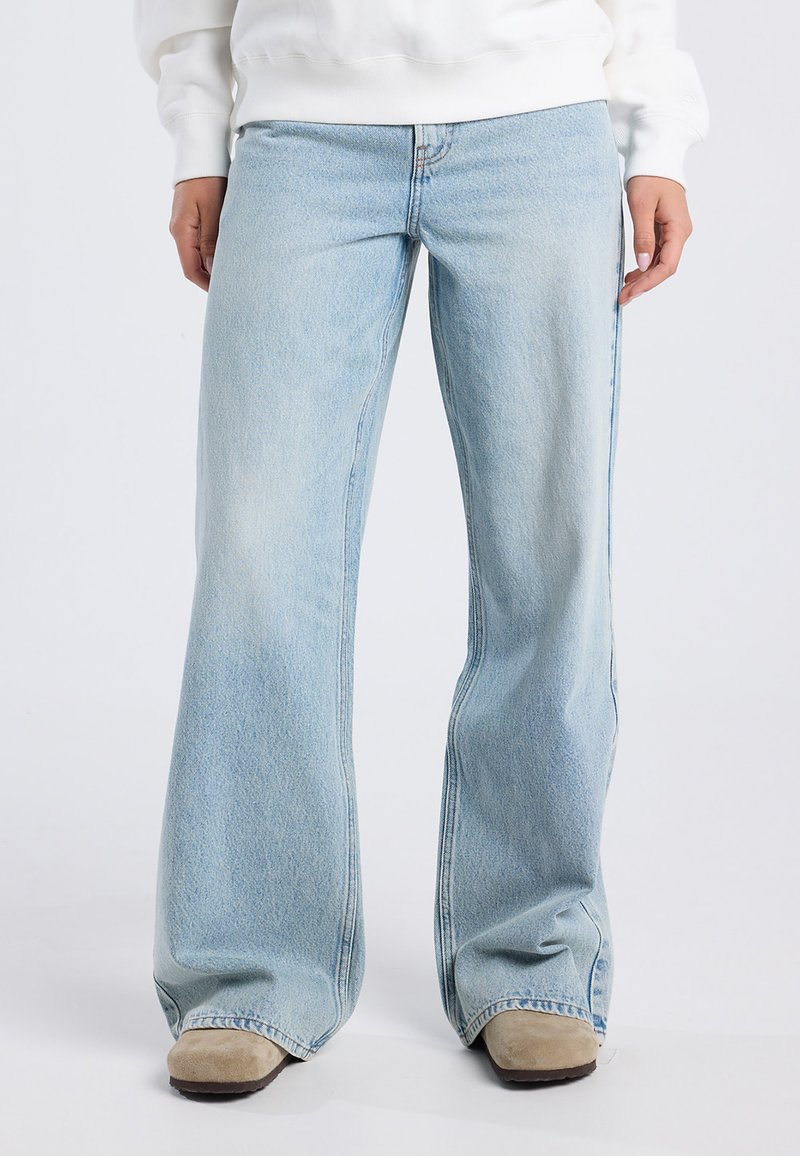 Light blue, flared jeans made of denim with a faded finish. Features subtle stitching details and wide leg openings, worn with beige footwear.