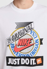 Una camiseta blanca de algodón con una impresión gráfica colorida. El diseño incluye "NIKE" en rojo, "JUST DO IT" en negrita negra y detalles de acento en amarillo y azul.