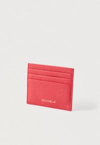 Rød tekstureret læder kortholder med tre kortslots og guld "COCCINELLE" logo på fronten mod en hvid baggrund.