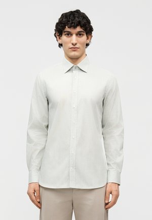 Νεαρός άνδρας με κατσαρά μαύρα μαλλιά, φορώντας ένα ανοιχτό γκρι πουκάμισο με λεπτές ρίγες και μπεζ παντελόνι, στέκεται μπροστά σε ένα απλό φόντο.