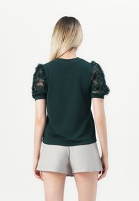 Mørkegrønn kortermet topp med gjennomsiktige, teksturerte pufferm. Designet har volanger på ermene og en klassisk halslinje.