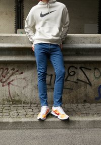 Vaaleanharmaa Nike-huppari mustalla logolla, siniset farkut ja valkoiset tennarit, joissa oransseja ja punaisia yksityiskohtia, seisoo mukulakivikadulla.