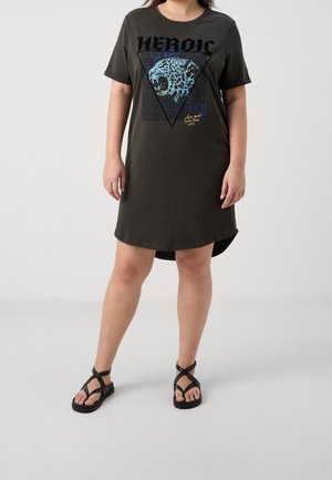 Personne portant une robe t-shirt oversize foncée avec un dessin léopard rugissant et un texte "HEROIC", associée à des sandales noires à lanières.