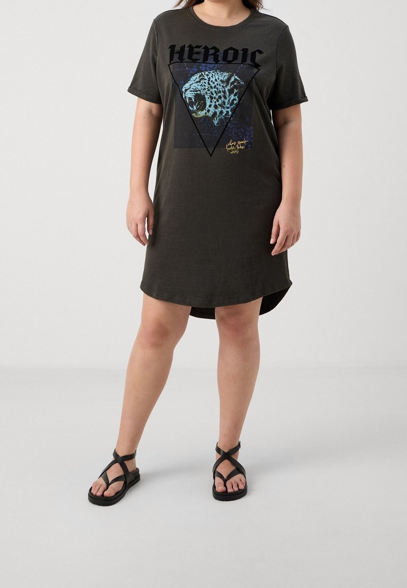 Personne portant une robe t-shirt oversize foncée avec un dessin léopard rugissant et un texte "HEROIC", associée à des sandales noires à lanières.