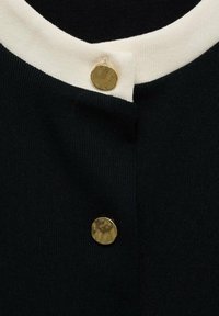 Abbigliamento in maglia nera con un colletto a coste color crema e due bottoni metallici dorati al collo. Texture liscia con un design aderente.