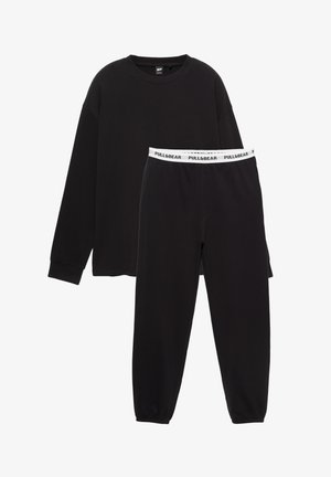 Set de îmbrăcăminte relaxată neagră, format dintr-un top cu mânecă lungă și pantaloni conici. Topul are un decolteu rotund; pantalonii au o talie albă cu logo.