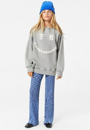 Grå sweatshirt med vit texturering, kombinerat med blå mönstrade utsvängda byxor. Modellen bär en blå stickad mössa. Svarta skor syns.