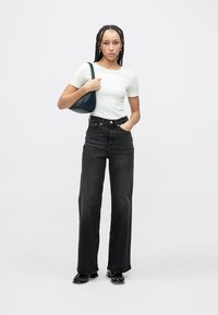 Bijela uska majica, crne traperice visokog struka, crne cipele i mala plava torbica. Model stoji u sredini na jednobojnoj pozadini.