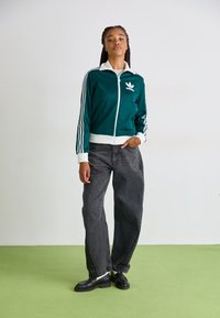 Casaco de treino verde-água com detalhes brancos e três riscas laterais, combinado com jeans largos pretos e sapatos pretos num chão verde.