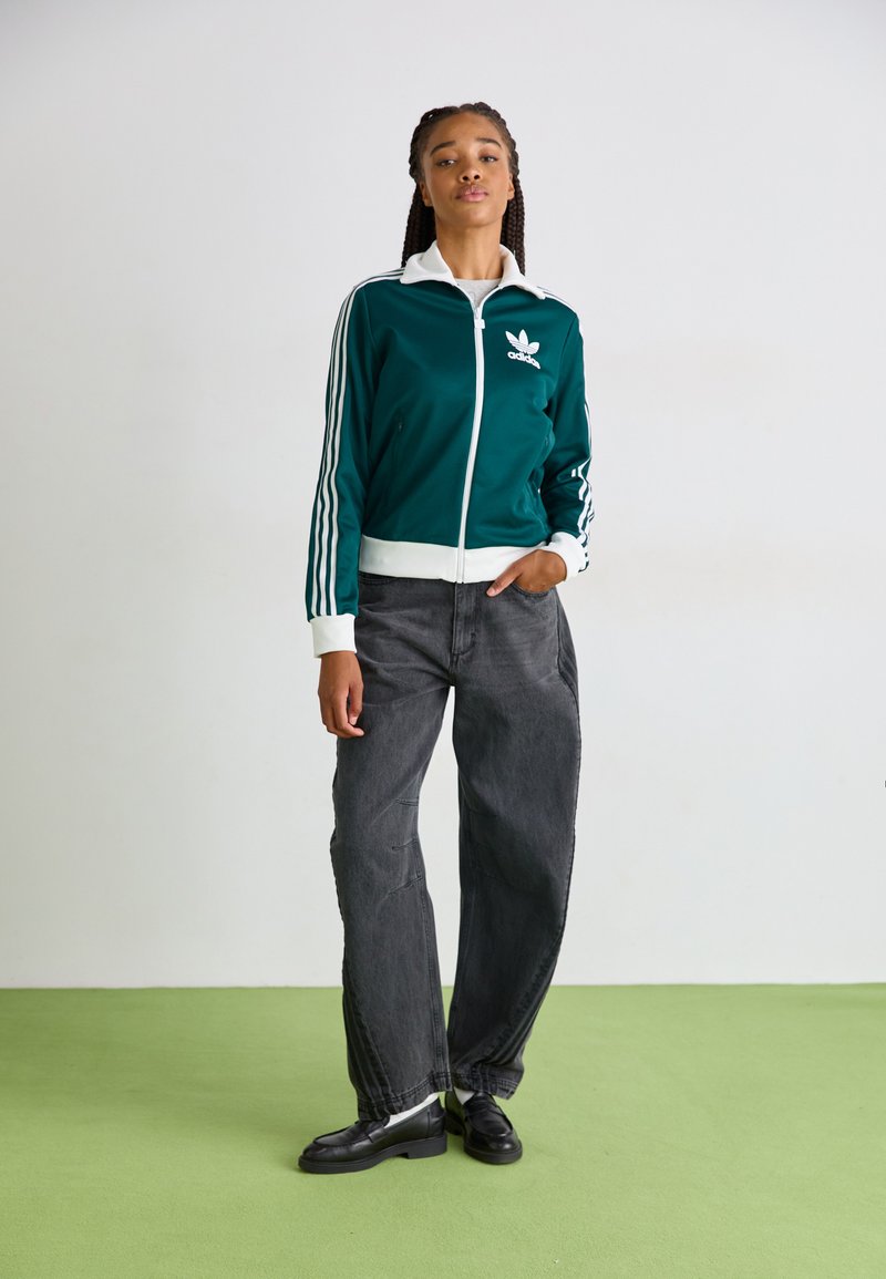 Teal Trainingsjacke mit weißen Akzenten und drei Seitenstreifen, kombiniert mit lockeren schwarzen Jeans und schwarzen Schuhen auf einem grünen Boden.