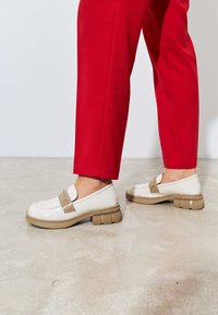 Valkoiset patenttivetävät loaferit, joissa on beige hihna, paksu urittu pohja ja accenttikoristelu, yhdistettyinä punaisiin housuihin neutraaliolla.