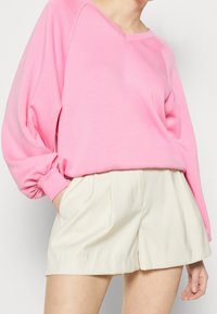 Różowa bluza z długim rękawem, dekoltem w szpic i bufkami, zestawiona z kremowymi, plisowanymi szortami. Miękka faktura tkaniny.