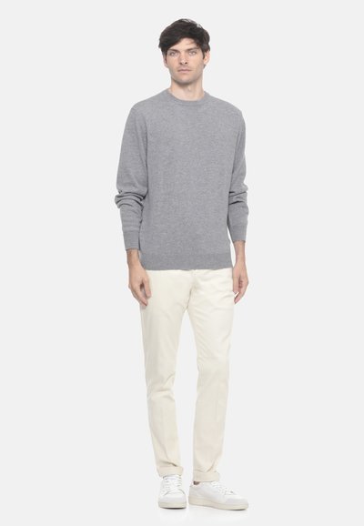 Uomo in piedi che indossa un maglione grigio a maniche lunghe, pantaloni avorio e sneakers bianche su uno sfondo chiaro e uniforme.