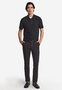 Svart polo shirt med en treknapps placket, i kombination med mörka byxor och svarta loafers. Slät tyg och figursydd design. Händerna i fickorna.