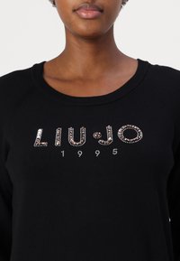 Gros plan d'une personne portant un pull noir avec "LIU·JO" et "1995" décorés de perles brillantes sur la poitrine.