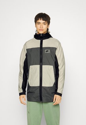 Giacca a vento Nike con cappuccio, design color-block con sezioni beige, grigie e nere, chiusura con zip e due tasche frontali.