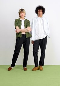 Two models stand against a white background. One wears a green button-up shirt and black pants; the other has a striped button-up over a white t-shirt. Both wear brown shoes.