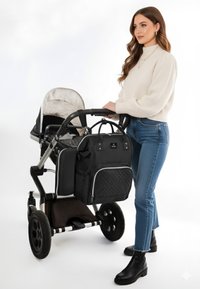 Sort barnevogn med en blød grå indvendig, parret med en kviltet sort pusletaske. Kvinde i en cremefarvet sweater og blå jeans står ved siden af den.