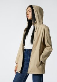 Beige waterdichte jas met een capuchon, voorzien van een ritsluiting, drukknoopsluitingen en zijzakken. Afgewerkt met een gladde textuur en een relaxte pasvorm.