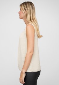 Hellbeige ärmelloses Oberteil aus strukturiertem Stoff, mit Rundhalsausschnitt und seitlichen Schlitzen, getragen über schwarzen Leggings.