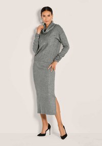 Frau posiert in einem grauen Strickpullover mit Zopfmuster und einem passenden gestrickten Midirock mit seitlichem Schlitz, trägt schwarze High Heels.