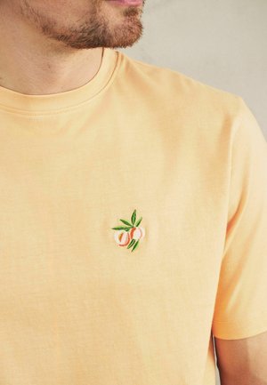 Mężczyzna ubrany w jasno-pomarańczową koszulkę z małym haftowanym wzorem brzoskwini na lewej stronie klatki piersiowej na jednolitym tle.