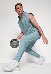 Lyseblå ærmeløs hoodie med kængurulomme, matchende taperede bukser og hvide sneakers med guldaccenter, som holder en sort og grå basketball.