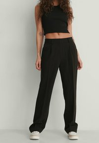 Vrouw in een zwart mouwloos cropped top, hoge, losse zwarte broek, en witte sneakers, staand tegen een effen lichte achtergrond.