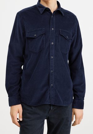 Mann trägt ein langärmeliges marineblaues Kordhemd mit geknöpften Brusttaschen und dunklen Hosen, sichtbarer Oberkörper, entspannte Arme.