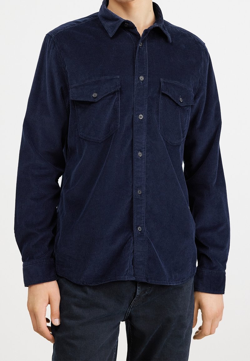 Homme portant une chemise en velours côtelé bleu marine à manches longues avec poches poitrine boutonnées et pantalon foncé, torse visible, bras détendus.