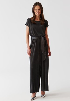 Zwarte geribbelde jumpsuit met korte mouwen, een aangesloten tailleband en een wijd uitlopende pijp. G gedragen met zilveren hoge hakken. Soepele textuur overal.