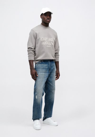 Sudadera gris con el logo "EMPORIO ARMANI", jeans azules desgastados y zapatillas blancas. El modelo lleva una gorra blanca. Conjunto simple y casual.