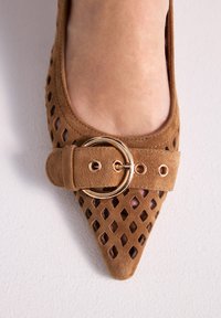 Zapato plano puntiagudo de ante marrón con recortes en forma de diamante y una correa con hebilla circular dorada sobre un pie desnudo.