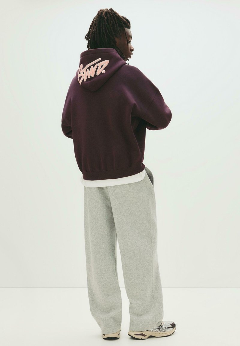 Felpa cappuccio bordeaux con logo scritto a mano sulla schiena, abbinata a pantaloni della tuta grigi larghi e scarpe da ginnastica, vista da dietro.