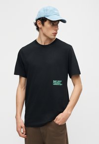 Νεαρός άνδρας που φοράει μαύρο μπλουζάκι με πράσινο κείμενο κοντά στη μέση και ανοιχτό μπλε καπέλο μπέιζμπολ, κοιτάζοντας προς τα δεξιά.