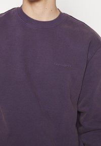 Fioletowa bluza z miękkiego materiału, z dekoltem w serek, ściągaczami przy mankietach i dyskretnym haftowanym logo Carhartt na piersi.