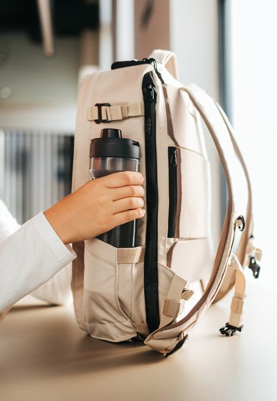 Mochila beige con múltiples compartimentos, que cuenta con un bolsillo lateral que sostiene una botella de agua negra. Tela duradera y correas ajustables.