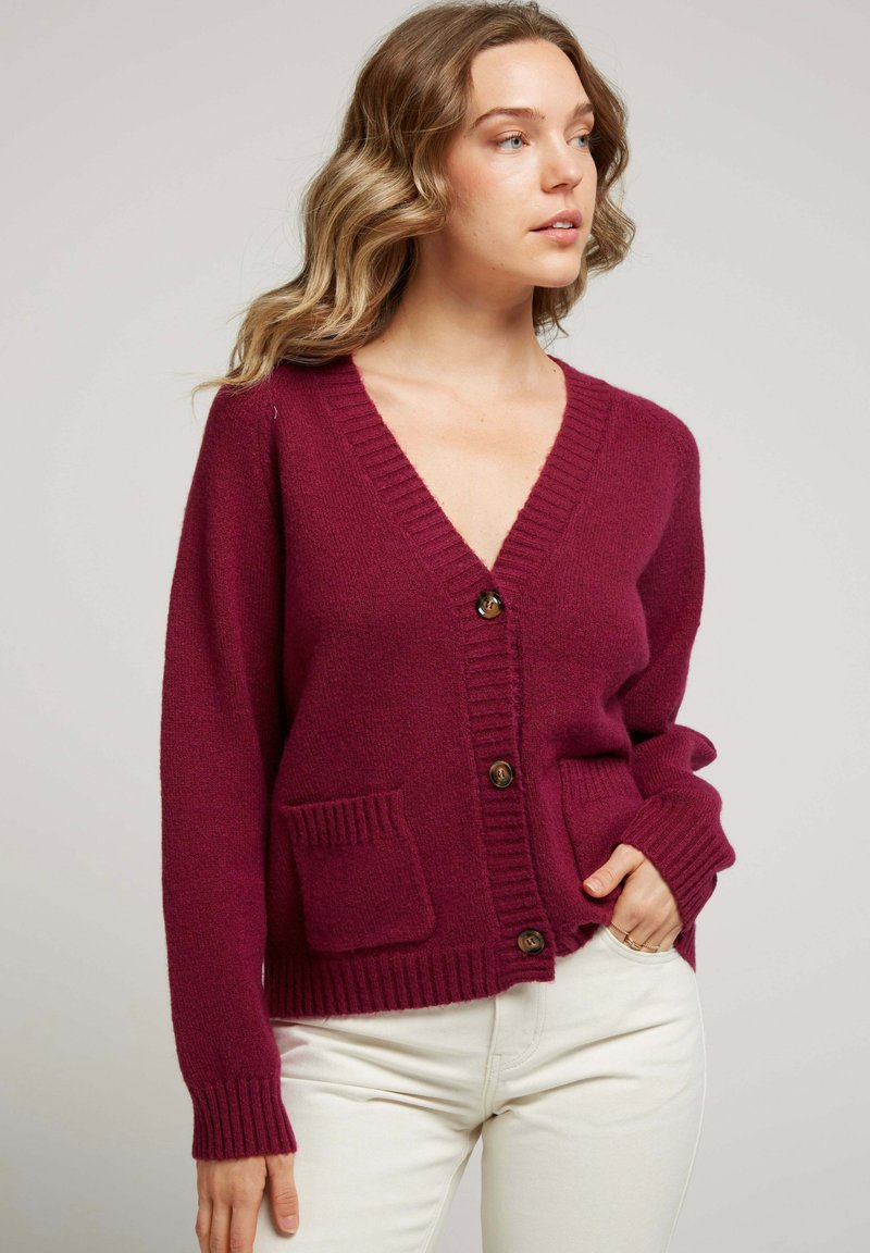 Cardigan en maille bordeaux avec un col en V, fermeture à boutons, poignets côtelés et deux poches avant. Texture douce avec une coupe décontractée.