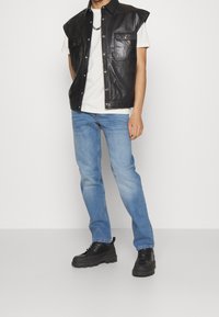 Černá kožená bezrukávová košile s předními kapsami, doplněná světle modrými džínami a černými robustními teniskami. Pod ní bílý tričko.